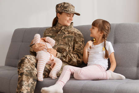 Horizontal shot of winsome soldier woman wearing camouflage uniform sitting on cough with two daughters, holding infant baby in hands and talking with elder kid.の写真素材