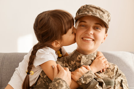 Indoor shot of woman soldier returning home from army, wearing camouflage uniform and cap, charming daughter hugging and kissing mother's cheek, missing mommy so much.の写真素材