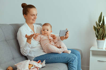 Positive adorable female wearing white shirt and jeans sitting on sofa with her infant daughter and having video call with friend or broadcasting livestream, spreading palm aside.の写真素材