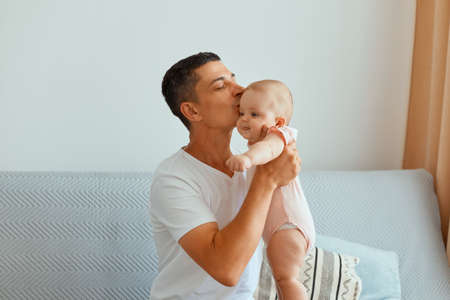 Indoor shot of caring handsome man wearing white t shirt sitting on sofa and kissing her charming cute little toddler daughter, spending his weekend with child.の写真素材