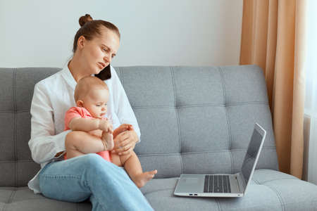 Indoor shot of sad upset woman with hair bun wearing white shirt and jeans, having sorrow expression, has problems with freelance work, sitting on sofa with infant baby daughter.の写真素材