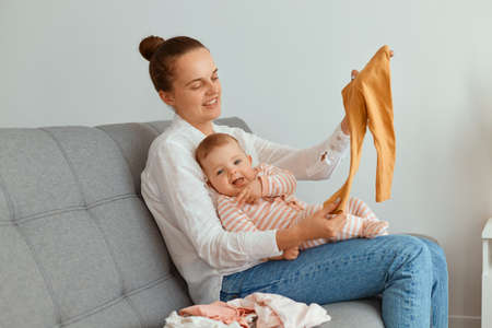 Portrait of smiling young adult mother sitting on sofa with her toddler daughter, holding baby's...