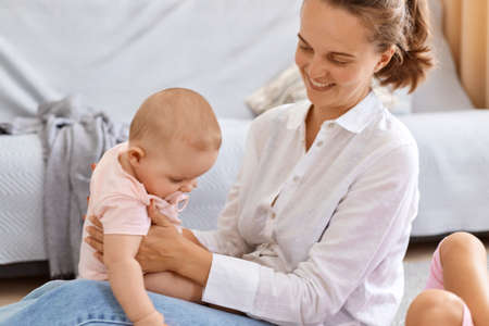 Indoor shot of smiling happy woman with dark hair woman playing with her infant daughter at home,...