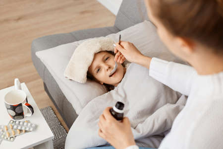 Portrait of little sick girl with wet towel on her forehead lying on sofa, mother giving her spoon with syrup, treating her lovely child from influenza or virus.の写真素材