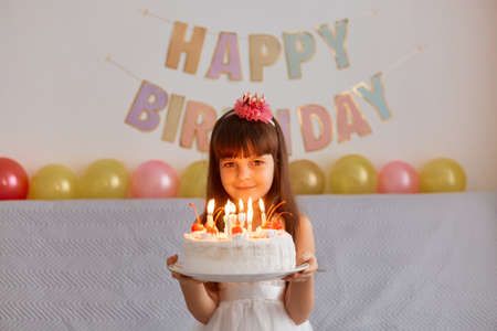 Portrait of happy smiling little girl standing holding cake with candles, celebrating birthday, being ready to make wish and blowing, posing indoor with festive inscription and balloons on backgroundの写真素材
