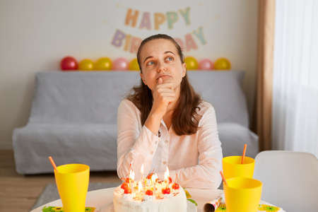 Portrait of sad lonely young adult thoughtful woman sitting alone at birthday table with cake, holding chin and looking away, having pensive expressing, thinking about birthday wish.の写真素材