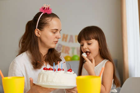 Horizontal shot of mother with daughter celebrating birthday, little girl licking finger tasting delicious festive dessert, home party, happy holiday with family.の写真素材