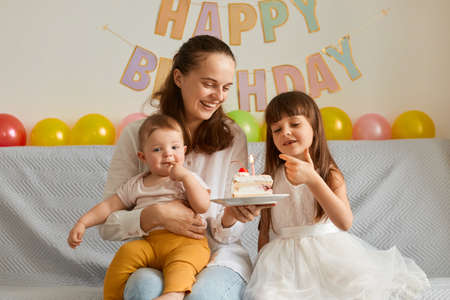 Image of happy smiling family, woman with her daughters, sitting on sofa with birthday cake,...