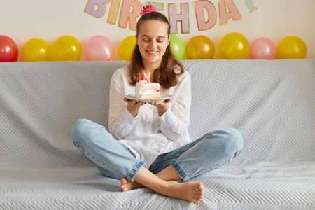 Portrait of happy positive young woman wearing white shirt and jeans sitting on sofa with crossed legs, holding cake, being ready to blowing candle, festive decoration on background.の写真素材