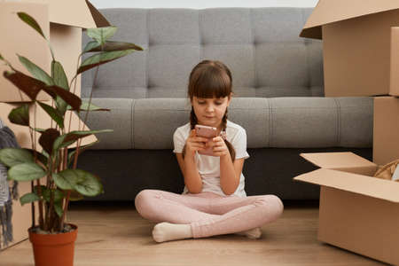 Portrait of concentrated little girl with braids wearing casual style clothing sitting on a floor near cough, playing game on mobile phone, surrounded with packages during relocating.の写真素材