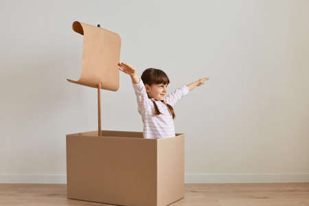 Indoor shot of excited little girl sitting in cardboard box with paper flag, playing ship capitan, pretending she is in the sea, spreading her hands aside like flying, enjoying her game.の写真素材