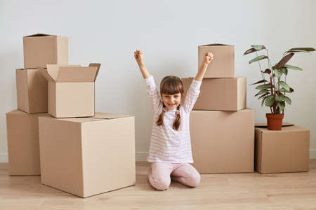 Indoor shot of happy excited little dark haired girl sitting on floor surrounded with cardboard boxes with personal belongings, being happy to move to a new apartment, celebrating, raised arms.の写真素材