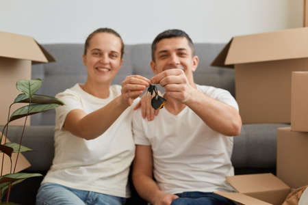 Indoor shot of couple holding key ring to their new house, looking at camera with charming smile, relocating to a new house, buying or renting new apartment.の写真素材