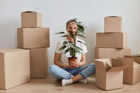 Horizontal shot of woman wearing white T-shirt sitting on the floor near cardboard boxes with...