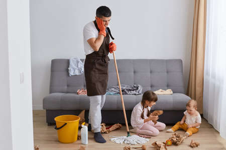 Full length portrait of upset Caucasian man wearing brown apron cleaning apartment, washing floor with mop, keeping hand on his cheek, being sad of disorder made of his children.の写真素材