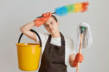 Portrait of tired woman wearing brown apron and white t shirt cleaning her flat, holding mop, ppduster and bucket, keeping hand on her forehead, looking exhausted of household chores.の写真素材
