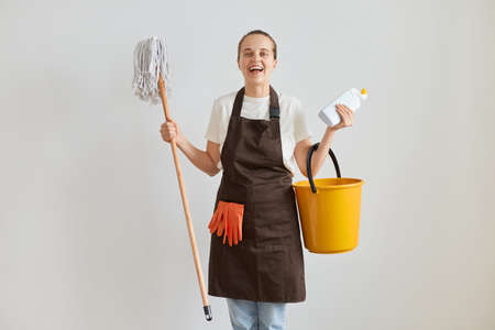 Extremely happy excited woman wearing brown apron and jeans washing floor at home, standing with...