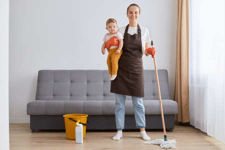 Indoor shot of positive optimistic woman wearing brown apron, jeans and white t shirt cleaning her...