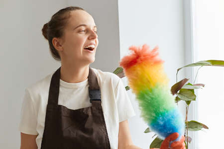Portrait of excited happy smiling woman wearing brown apron and white t shirt cleaning her flat, having fun while doing housework, singing with multicolored ppduster.の写真素材