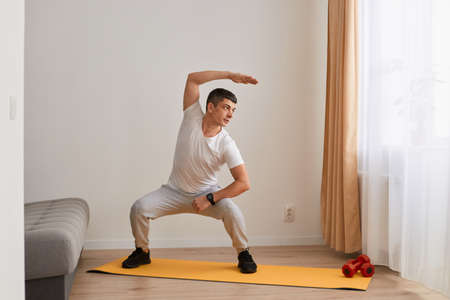 Strong man wearing sportswear and sneakers standing squatting on mat practicing yoga, bending to...