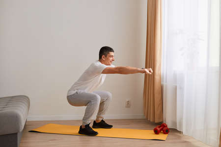 Side view portrait of athletic man wearing sportswear doing squat, lower body sport exercise, warming up and training muscles, keeps arms outstretched, looking ahead.の写真素材