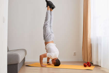 Indoor shot of sporty man with dark hair standing upside down on the head near the sofa, yoga and...