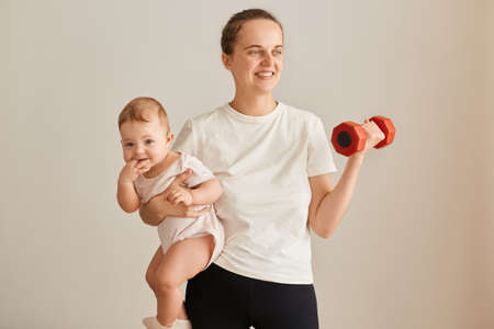 Indoor shot of attractive athletic woman wearing white t shirt holding baby daughter in hands and training arms with raising dumbbell, mother doing sport exercises against while wall at home.の写真素材