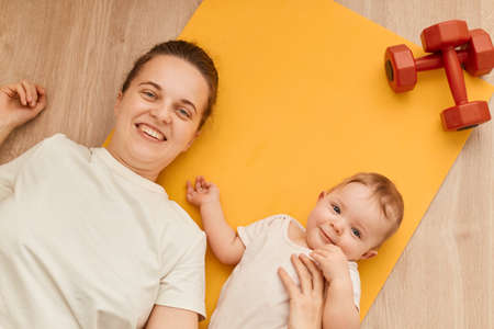 Top view of smiling woman lying on yellow yoga mat with her infant daughter near red dumbbells, looking at camera, expressing happiness, fitness at home.の写真素材