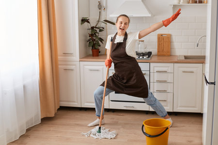 Portrait of excited extremely happy housewife wearing white t shirt, jeans and brown apron, washing floor with mop and dancing, having fun while cleaning her apartment.の写真素材
