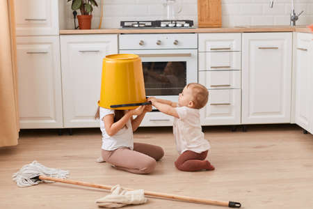 A portrait of two little girls sitting on the kitchen playing with cleaning supplies, the older sister sitting with a bucket on her head, cleaning the house.の写真素材