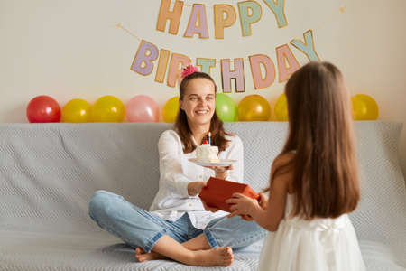 Portrait of happy woman celebrating her birthday, little cute daughter posing backwards to camera and giving mother her gift in red box, festive decoration on background.の写真素材
