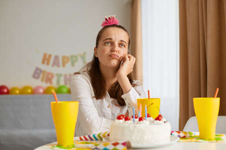 Image of bored attractive young woman wearing white shirt celebrating birthday, sitting alone at table with cake, keeping hand under chin, looking away, dreaming.の写真素材