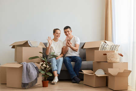 Horizontal shot of smiling young family buying new flat and posing surrounded with boxes with personal pile, sitting on sofa and having video call, waving hands to phone camera.の写真素材