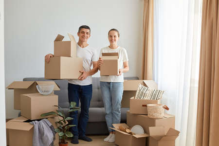 Full length portrait of woman and man, young couple wearing white t shirts standing with cardboard boxes, relocating to a new house, unpacking personal pile, looking smiling to camera.の写真素材