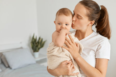Indoor shot of young adult attractive woman holding daughter wrapped in towel after bathroom, mother kissing her lovely baby girl, expressing love and gentle.の写真素材