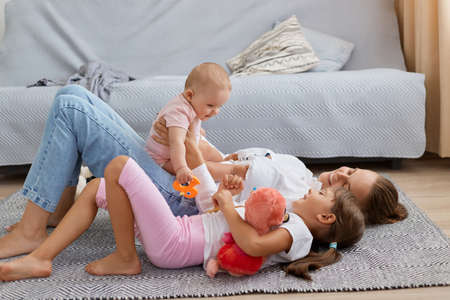 Side view portrait of smiling optimistic woman lying on floor with her daughters, baby kid sitting on mom's belly, elder child lying near mommy, family spending happy time together.の写真素材