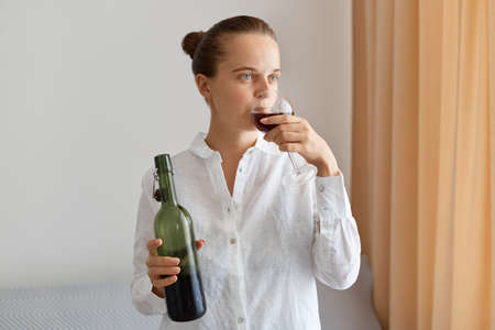 Indoor shot of young adult Caucasian woman with hair bun wearing white shirt holding bottle of wine and drinking beverage from glass, looking away, delicious taste.の写真素材