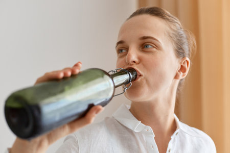 Closeup portrait of attractive woman with perfect skin wearing white drinking shirt of wine from bottle, female alcoholic enjoying tasty drink, posing with white wall and curtain of background.の写真素材