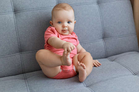 Horizontal shot of cute charming aby girl wearing pink bodysuit, looking at camera, playing alone, touching her leg.の写真素材