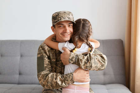 Portrait of smiling soldier man in camouflage hugging his daughter and looking at camera, sitting on cough at home, arrived home from army and being happy to be with his child.の写真素材