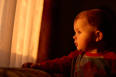 Indoor side view shot of charming infant baby looking at window, sun light shining pass through the window, beautiful sunset and cute toddler kid.の写真素材