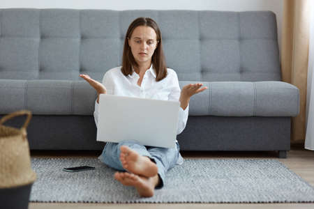 Indoor shot of puzzled helpless young adult woman wearing white shirt and jeans sitting on floor near cough and working on laptop, looking at display with spread arms and confused expression.の写真素材
