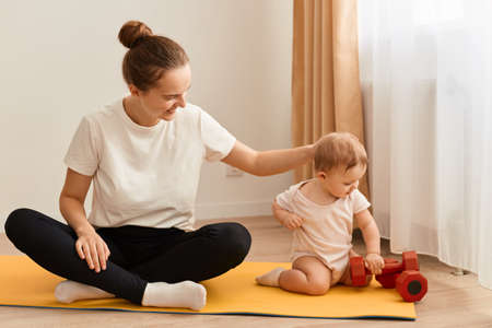 Image of woman with hair bun wearing white t shirt and black leggins doing sport exercises with baby...