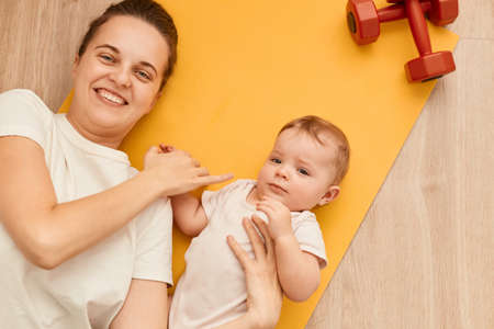 Top view of happy attractive woman lying on yellow yoga mat with her toddler daughter near red...