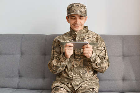 Indoor shot of smiling delighted military man wearing camouflage uniform sitting on sofa, soldier holding smart phone, playing online game while having time for leisure.の写真素材