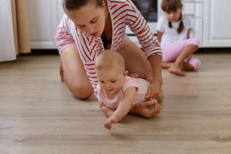 Horizontal shot of young adult woman wearing striped casual style shirt sitting on floor in kitchen...