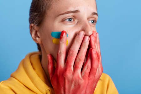 Portrait of Caucasian woman wearing yellow hoodie crying, covering mouth with palms with blood,...