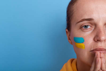 Closeup portrait of half of woman face wearing yellow hoodie and with flag of Ukraine on her cheek,...