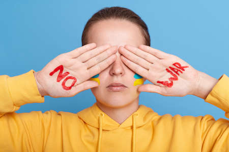 Portrait of unknown woman wearing yellow hoodie standing and covering her reyes with palms. Stop violence, sign on hand draw with red paint. don't want to see events in Ukraine.の写真素材