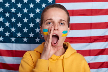 Woman wearing hoodie with flag of Ukraine on her cheek, yelling, screaming, posing isolated over flag of united states of america, Ukraine's war against Russia, assistance from allied countries.の写真素材